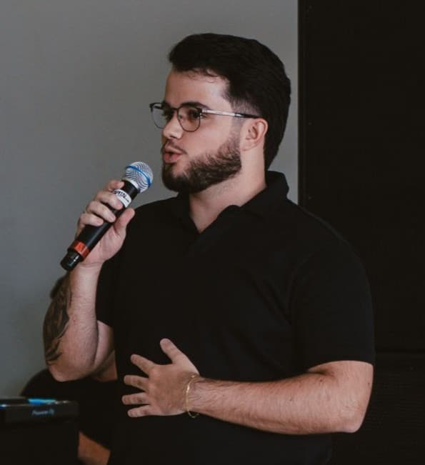Gustavo Gomes Balbino apresentando com microfone diante de uma plateia.